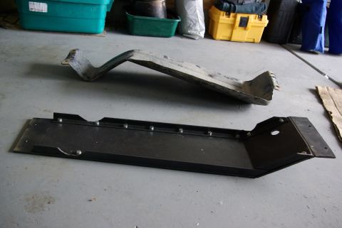 1995 Ford Bronco - Two long steel skid plate / crossmember pieces on a garage floor, a bare-metal stamped original standing on edge behind, and a heavier fabr…