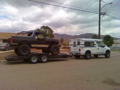 2000 Ford F-250 - A lifted black Ford Bronco is transported on a trailer by a white Ford F-250 pickup truck with a camper shell.