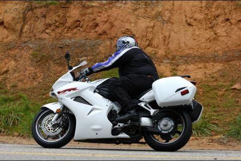 1985 Ford LTD Crown Victoria - A rider on a white motorcycle with saddlebags navigates a road with a rocky embankment in the background.