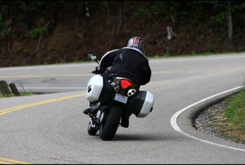 1985 Ford LTD Crown Victoria - Motorcycle with saddlebags navigates a curve on a paved road surrounded by trees.