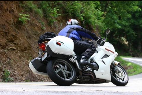 1985 Ford LTD Crown Victoria - Motorcyclist riding a white sport bike with saddlebags on a wooded road.