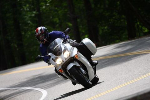 1985 Ford LTD Crown Victoria - A rider navigates a winding road on a white motorcycle, with trees lining the asphalt.