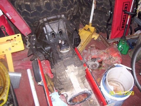 1978 Ford Bronco - A transmission part is shown on a red cart, surrounded by various tools and tires in a cluttered garage setting.