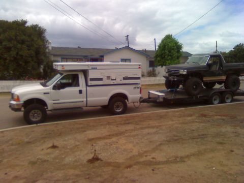 2000 Ford F-250 - A white pickup truck tows a trailer carrying a modified black pickup truck down a residential street.