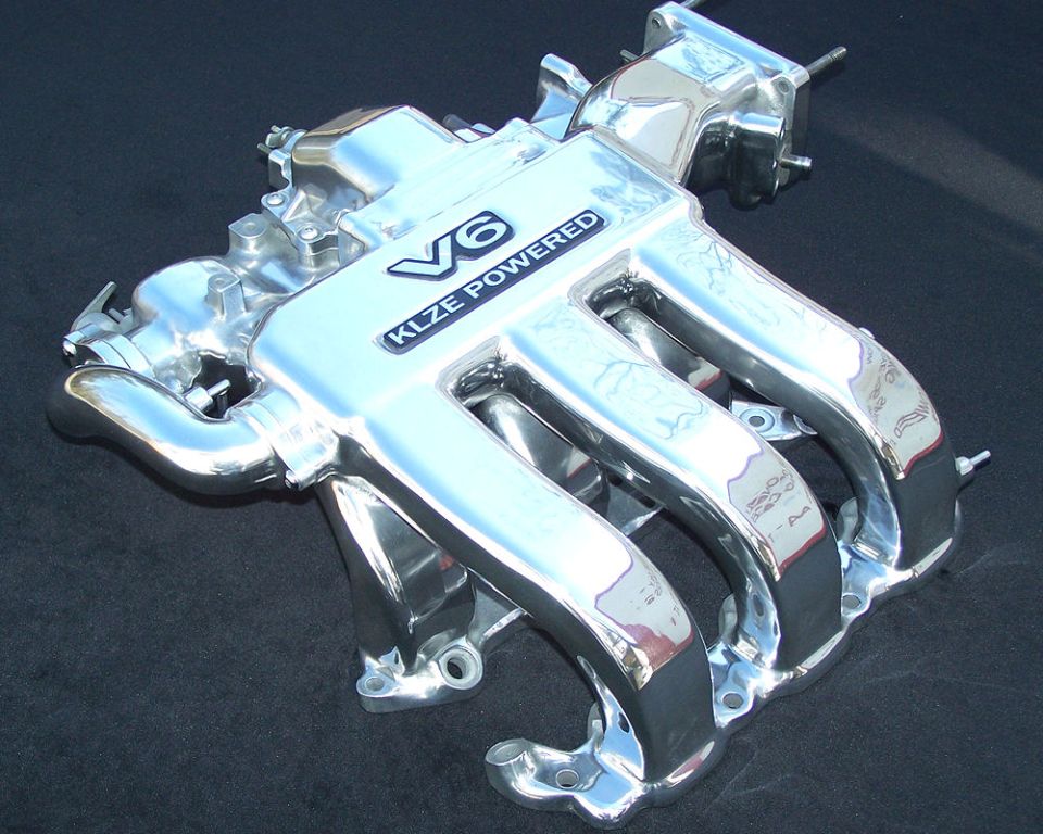 Polished chrome V6 intake manifold labeled KLZE Powered on a dark surface