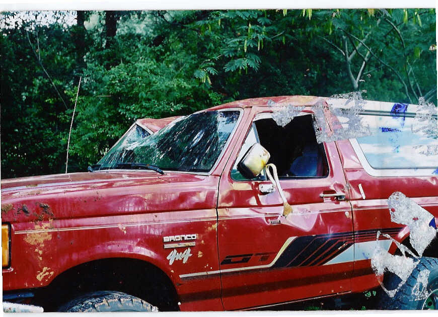 Side view of a rusty red Ford Bronco 4x4 SUV with cracked windshield, parked by trees
