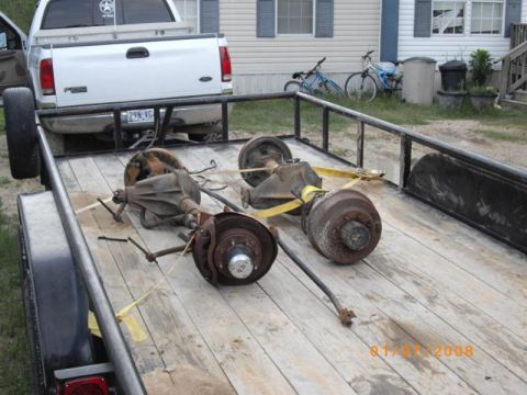1983 Ford Bronco - Trailer loaded with vehicle axles and brake drums, parked near a house with bicycles.