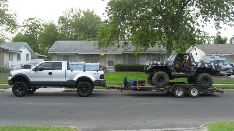 1979 Ford Bronco - A silver Ford F-150 tows a lifted, camo-wrapped Ford Bronco on a trailer down a residential street.