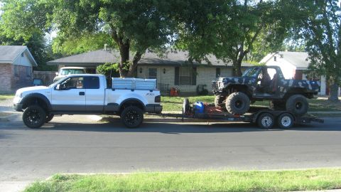 1979 Ford Bronco - A lifted pickup truck tows a trailer carrying a heavily modified, camouflage-painted SUV.