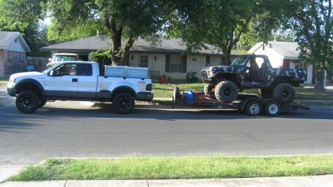 1979 Ford Bronco - A lifted, camouflaged Ford Bronco is transported on a trailer towed by a white pickup truck.