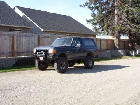 1988 Ford Bronco - Three-quarter front view of a lifted dark gray/blue full-size Ford Bronco sitting on a gravel driveway beside a cedar fence and a gray-side…
