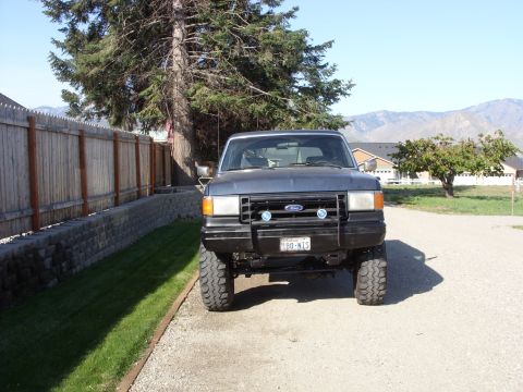 1988 Ford Bronco - Head-on view of a lifted dark blue Ford Bronco with a black tube grille guard carrying two round covered driving lights, Ford-badged grille…