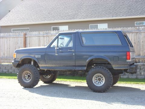 1988 Ford Bronco - Driver-side profile of a lifted dark blue full-size Ford Bronco with its removable hardtop in place, Bronco fender badge and body-side mold…