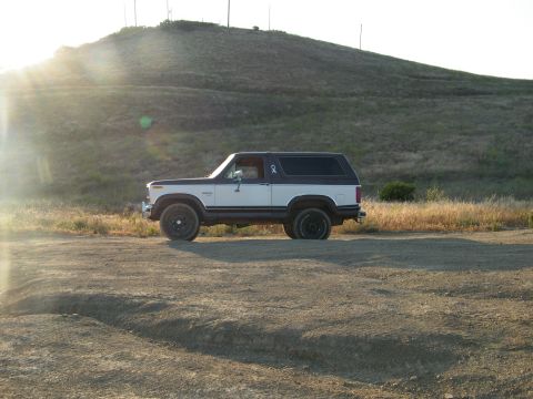 1982 Ford Bronco - A side view of a two-tone Ford Bronco sits on a dusty path under a bright sun.