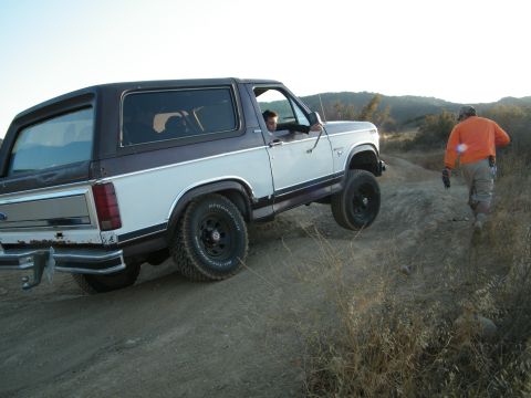 1982 Ford Bronco - A 1982 Ford Bronco navigates an off-road trail with a driver inside and a person walking nearby.