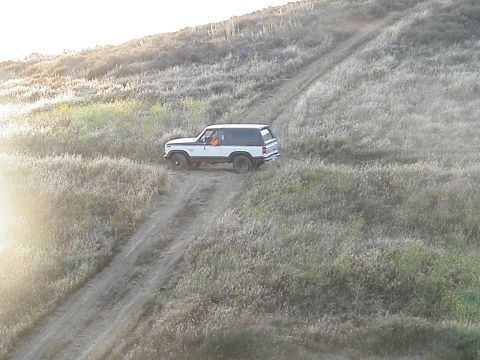 1982 Ford Bronco - A white SUV navigates a dirt road through a dry, grassy landscape under a bright sky.