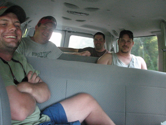 Four men smiling inside a van, one in foreground wearing green shirt and blue shorts