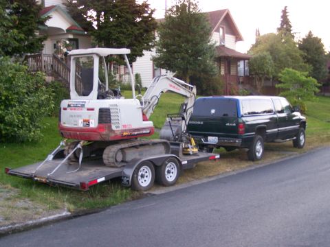 1988 Suzuki Samurai - A green Dodge Ram pickup truck tows an excavator on a trailer down a residential street.