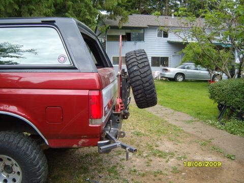 1989 Ford Bronco - The rear of a red SUV is shown with a spare tire mounted. A house and another car are visible in the background.