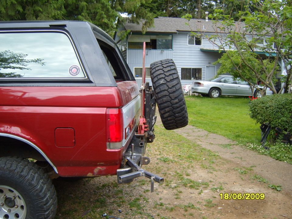 Red SUV's rear with spare tire on swing-away carrier. House and silver sedan in background