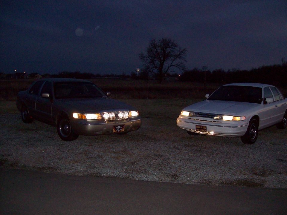 Two sedans, likely Ford Crown Victorias, parked outside at night. One is silver with extra lights on the grille, the other is white