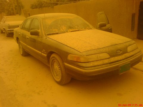1992 Ford Crown Victoria - Three-quarter front view of a dark aeroback Ford sedan parked on dirt beside a tan stucco wall, its hood, roof and windshield caked in dust…