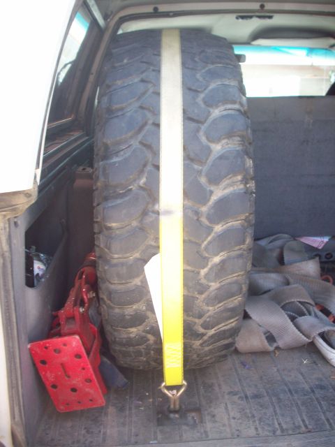 1995 Ford Bronco - A large spare tire is strapped down in the rear cargo area of a vehicle, next to a red jack.