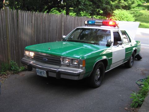 1991 Ford LTD Crown Victoria - Front three-quarter view of a green-and-white Ford police sedan with headlights on, parked in a driveway.