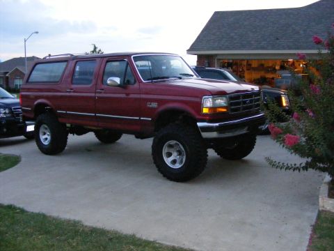 1993 Ford C-150 (Centurion Conversions) - A lifted red Ford Bronco SUV is parked in a driveway next to another vehicle, with a house and garage in the background.