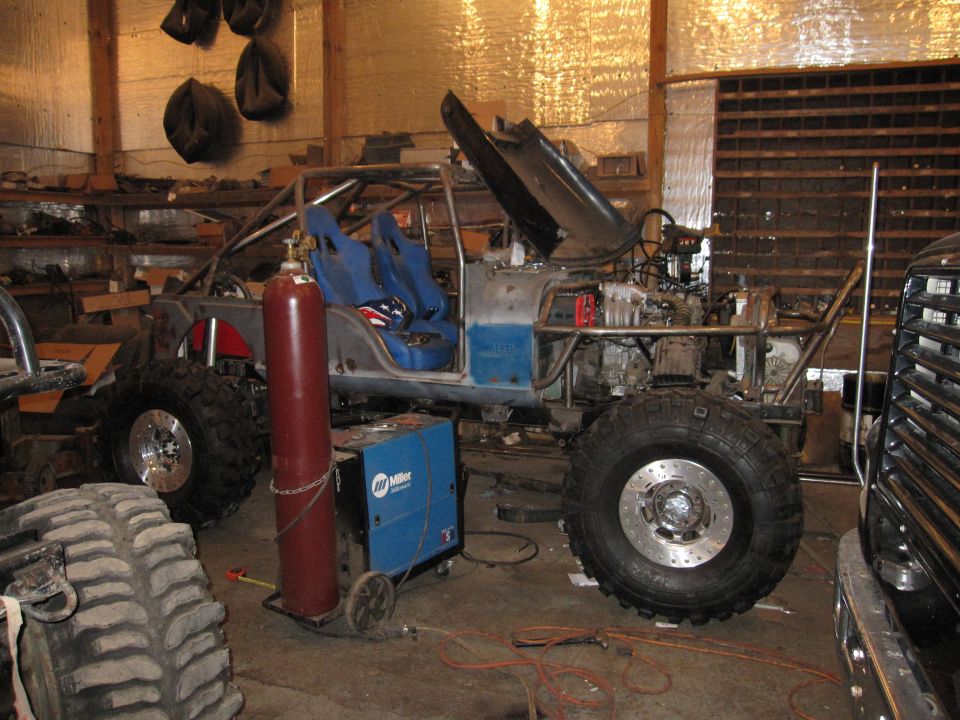 Custom off-road vehicle in garage with roll cage, racing seats, and exposed engine. Welding equipment nearby