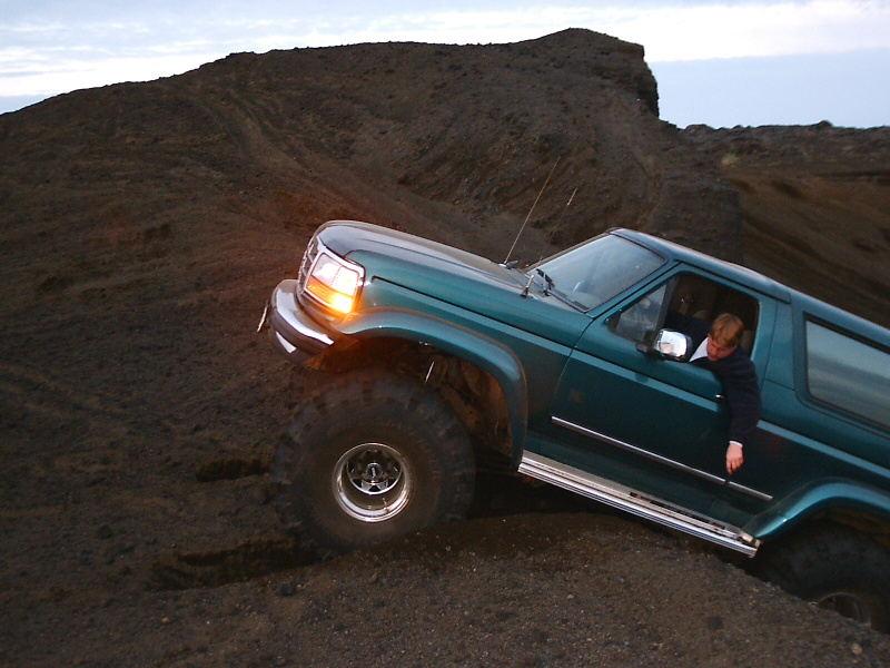 Teal lifted Ford Bronco on big off-road tires climbing a steep dirt hill at dusk