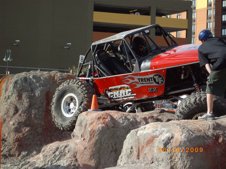 Red off-road buggy with roll cage on rocks, driver wearing helmet, person in black shirt and shorts nearby