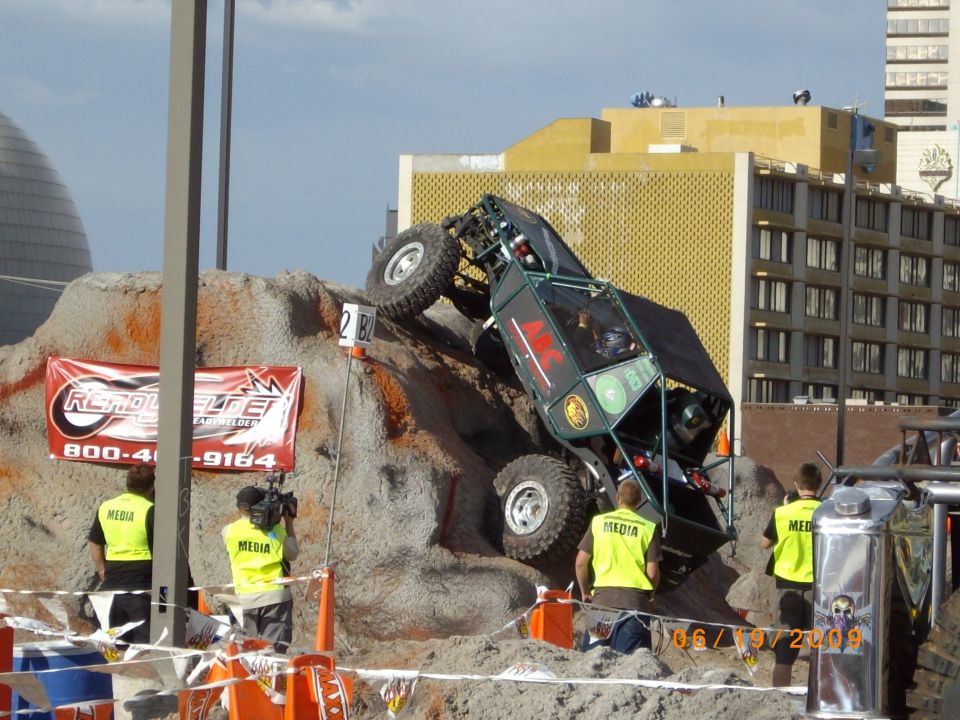 Off-road buggy climbing a dirt mound, media personnel present