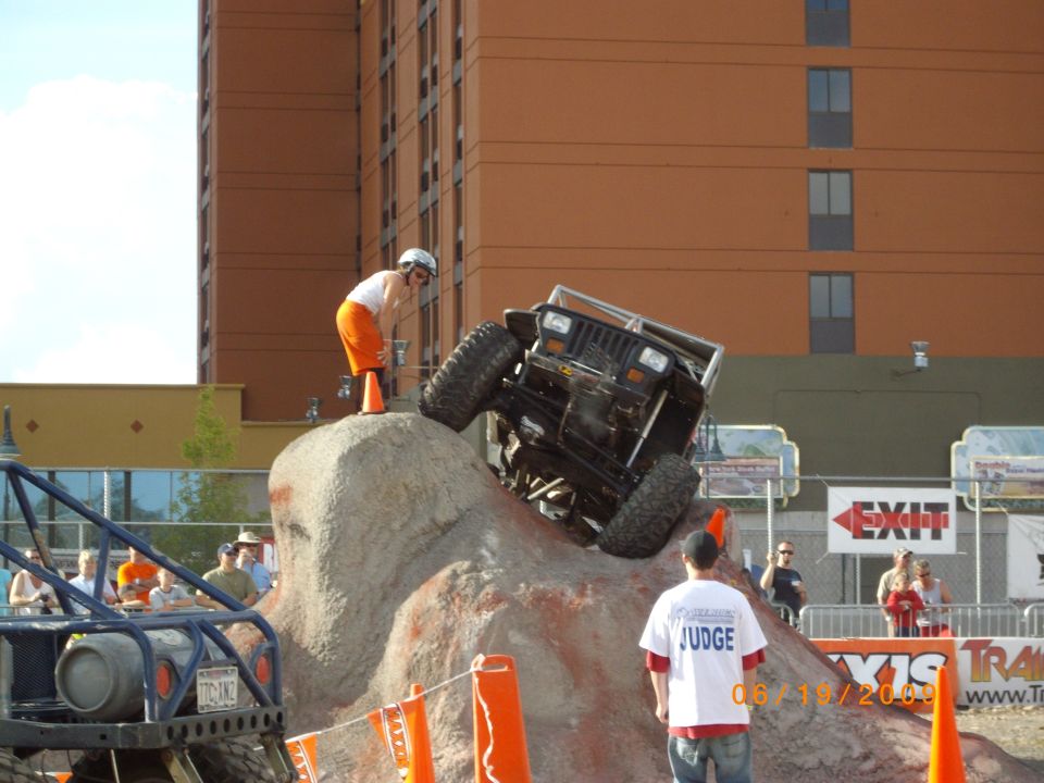 Black off-road vehicle tilted on a rock obstacle, driver wearing orange shorts and helmet, with a building in the background