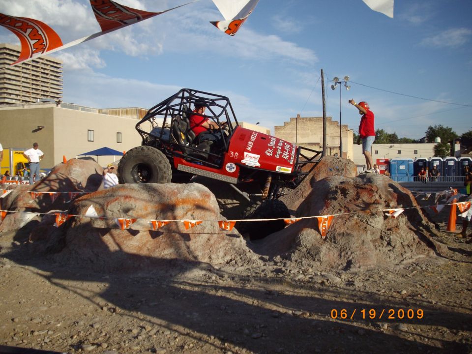 Red off-road buggy with a roll cage is climbing over rocks. A person in a red shirt stands on a rock, pointing