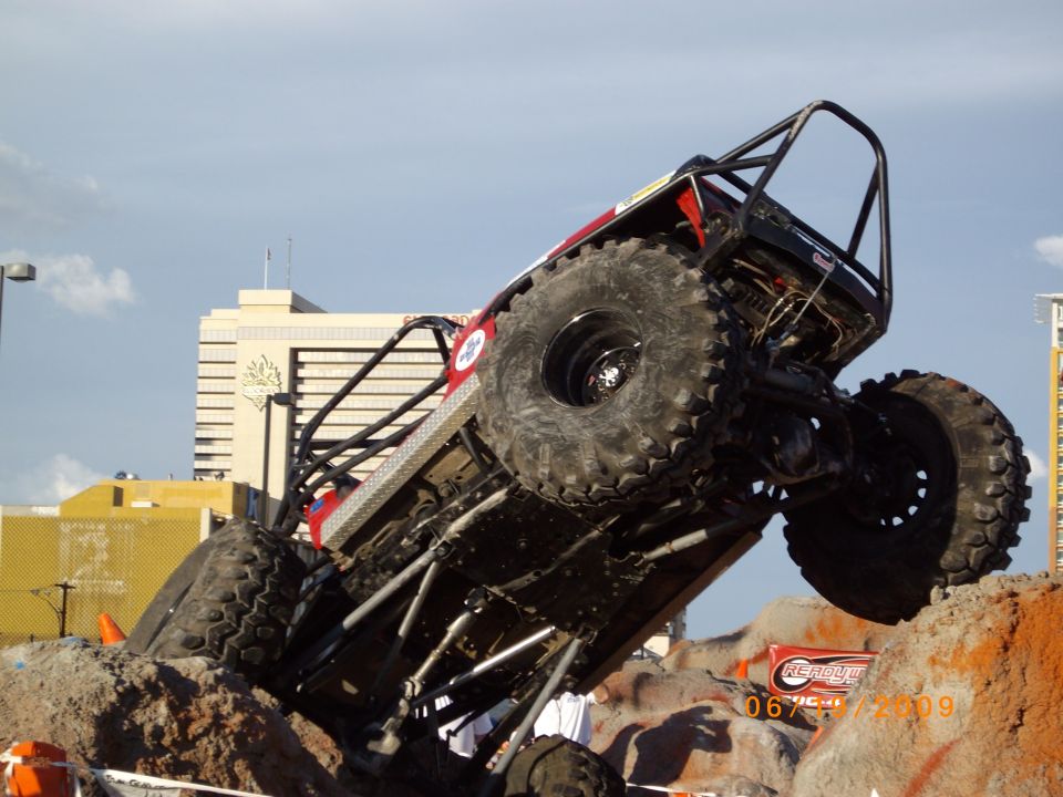 Off-road buggy with large tires climbing over rocks, with buildings in the background