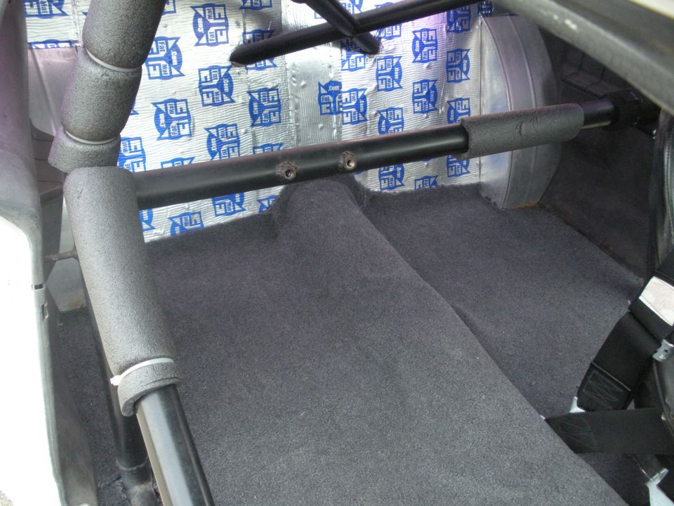 Interior view of a car showing a roll cage, grey carpet, and sound deadening material on the rear panel