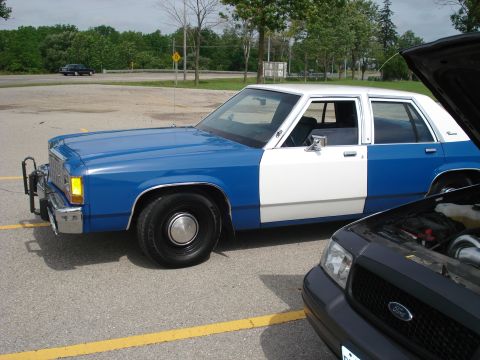 2000 Ford Crown Victoria - A two-tone Ford Crown Victoria is parked next to another vehicle with its hood open.