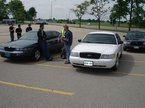 2000 Ford Crown Victoria - Two Ford Crown Victorias are parked in a lot, with a group of men standing near them.