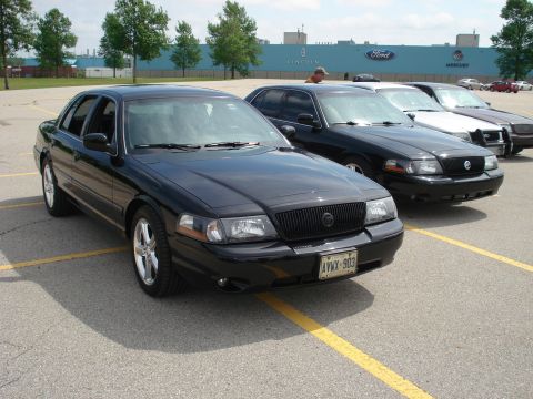 2000 Ford Crown Victoria - Several black sedans are parked in a lot with a factory in the background displaying Ford and Mercury logos.