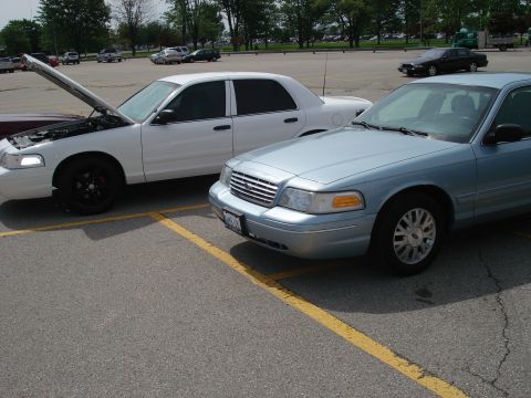 2000 Ford Crown Victoria - Two Ford Crown Victorias are parked in a lot. The white one has its hood open.