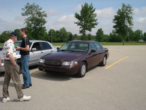 2000 Ford Crown Victoria - Two men stand near a maroon 2000 Ford Crown Victoria in a parking lot with another car visible.