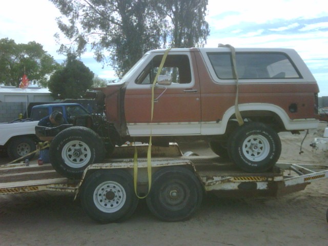 Red and white Bronco with front clip removed, strapped to a tandem-axle trailer, side view