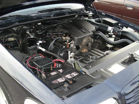 2000 Ford Crown Victoria - The engine bay of a 2000 Ford Crown Victoria, showing the V8 engine and battery. Another vehicle is parked behind it.