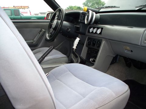 1988 Ford Mustang - The interior of a car shows light gray seats, a dashboard with gauges, and a manual shifter.