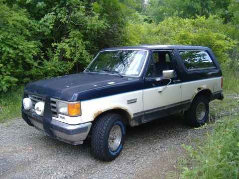 1988 Ford Bronco - A two-tone blue and white Ford Bronco with visible rust and aftermarket lights sits on a gravel road surrounded by greenery.