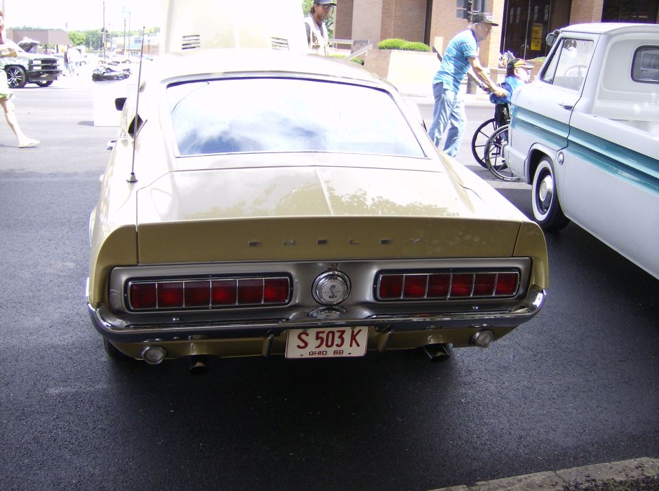 Rear view of a 1969 Ford Mustang Shelby GT500, gold with white stripes, parked next to a white pickup truck