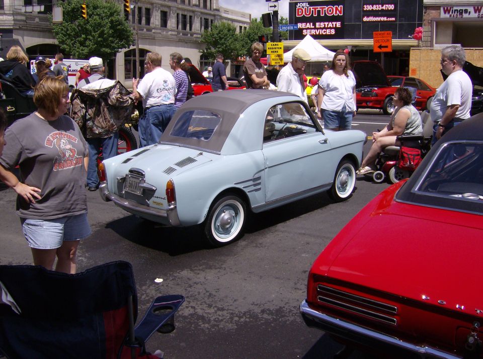 Light blue convertible car with a grey soft top, seen from the rear three-quarter angle