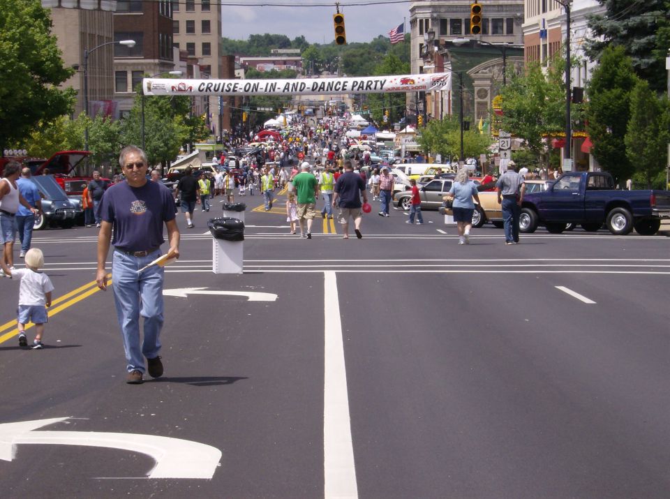 street filled with people and cars for a cruise-in event, banner overhead says 'cruise-on-in-and-dance party'