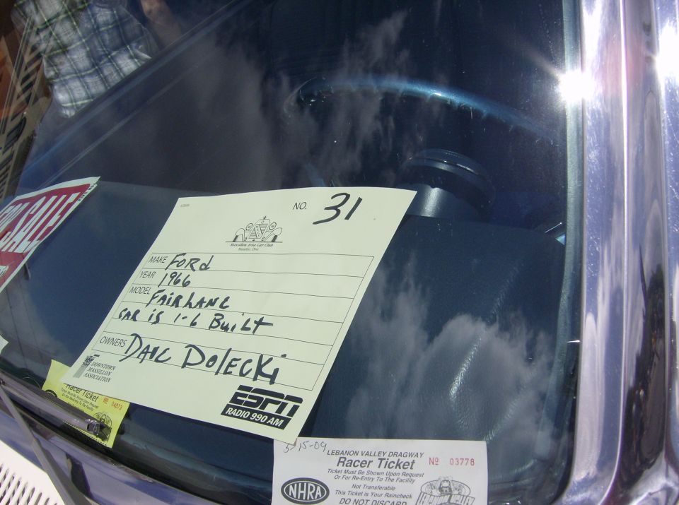 Car interior showing dashboard with a sign listing Make: Ford, Year: 1966, Model: Fairlane, Owner: Dale Dolecki. Also visible is a steering wheel and part of a plaid shirt